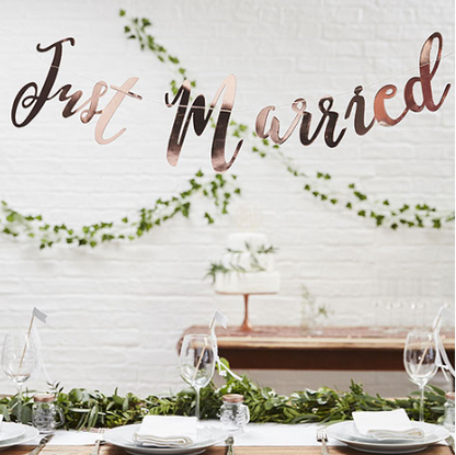 Decorative setup with 'Just Married' sign, tableware, and greenery against a white wall.