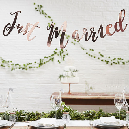Decorative setup with 'Just Married' sign, tableware, and greenery against a white wall.