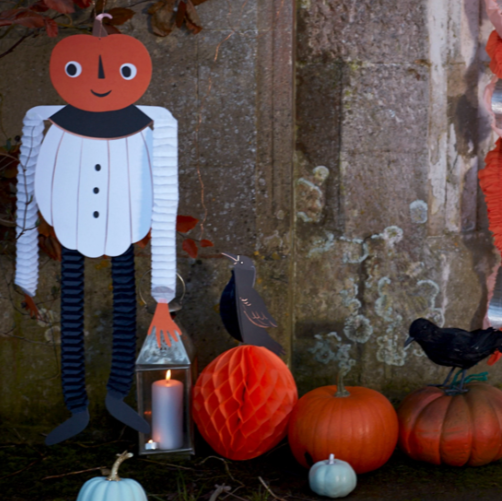 Halloween-themed decoration with a paper pumpkin character, lantern, and pumpkins against a textured wall.